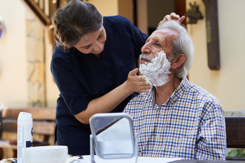 Professional helpful caregiver at nursing home. Health visitor and a senior man during home visit. Young caregiver in uniform helping to elderly man for shave his face