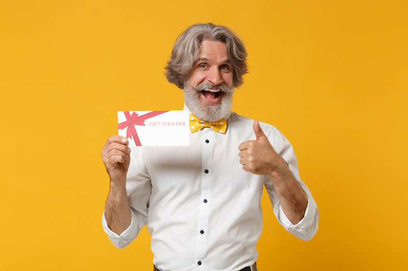 Funny elderly gray-haired mustache bearded man in white shirt bow tie isolated on yellow orange background. People lifestyle concept. Mock up copy space. Holding gift certificate, showing thumb up