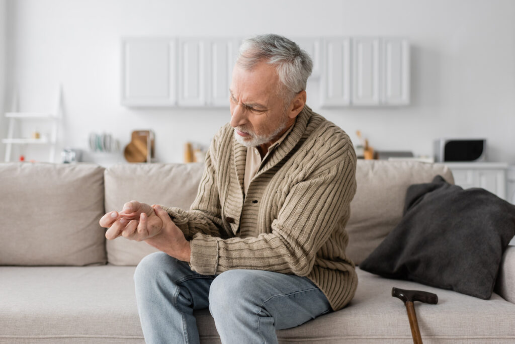 aged man with parkinson syndrome suffering from tremor in hands while sitting on sofa near walking cane,stock image