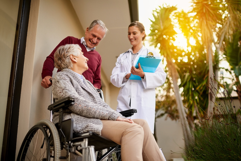 Senior, doctor and person with disability in retirement home for health, support or help for patient. Elderly, man or pushing woman with wheelchair in care facility, caregiver with report for medical.