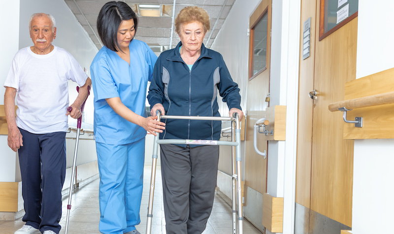 Asian doctor helping elder woman with walker and man in hospital aisle.