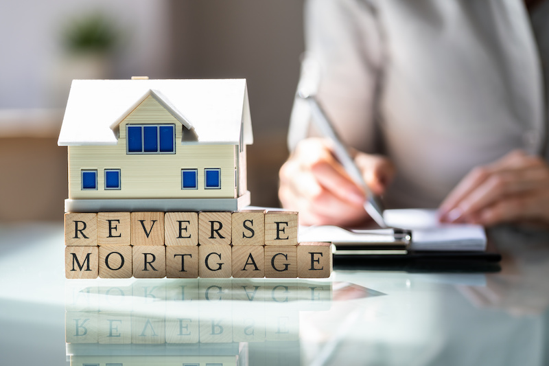 House Model Over Reverse Mortgage Blocks In Front Of Businesswoman Writing On Paper Over Desk