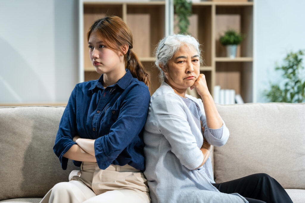 A mature mom and a young Asian woman, mother and daughter, sit together on a sofa in their living room, dealing with an argument and conflict, trying to resolve their differences at home.