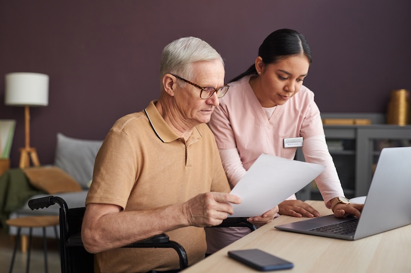 Young social worker in uniform helping senior man to pay bills online using laptop at table in the room