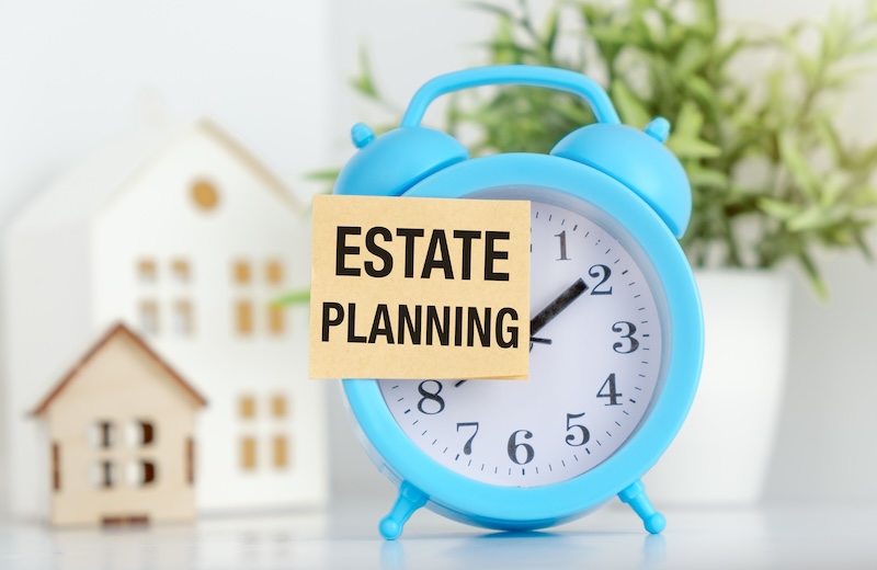 A blue alarm clock with a white paper that says Estate Planning on it. The clock is set to the time of 10:30