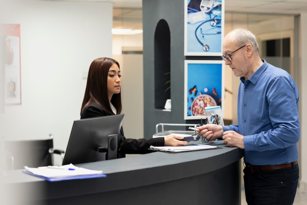 Female receptionist sits at clinic front desk, helping elderly male patient make payment after medical consultation. Senior man uses credit card on POS terminal as staff member provides assistance. SSUCv3H4sIAAAAAAAACmxUUW+kOAz+K4jnUiUhCTCvtyft0+6qutNJV06rEJKSLRAUQqtR1f9+jmFGM9I+QRz7s/19jj/yTq1O56eP3I3jtsagovNzfiIPeTBzbwL+voVSEvxTDj+md9EHp0Y8dSrqYVaTyU/zNo6fD/kaVdxWswIwnLSK5gXc8XzJ+PyR7Pkpz8F969LP58NvbP9B0hcz63PCArBgRqMQ+jknqrK6a2Rhre0KLuu6aKTgRV1WVNG6F5pRgJKCVRU1tuC9qgvONbj1ZVf0urQdtcqyyoAbV9Zy05jCdAx8O1EVTUVF0WjwgCBrbAVulPASAuqiZDUveMrcaVsXgglVdkI3tBE5VP36Hk2YsGVrJjUCO885lFc16VarTQMRCrh+ziWlNVqD0WZJCrg17heCkXSxmtn5kE27P6CIGlFGNyOZeVVTWaIpJHEyrUKPCEQwnuw2+DlmvVlfk5k1gjSSI8jikUzKZF1VyTCYcXHzyx5eEnSaAFWrMdN+XrcxHmMCeaF0bHfw6+JimgiIEoLJZDQjjNB4RlvFJE02mLF5VfoCICVrsMUbigS5SzkY/botd9kWKMDMB0elRNu7chHKzoL3EzoLvre+qPN0OFNSlw02pNYVSFazNnd97kTfFTx5aGKvlbEaSYZAaxGvqkE8guKpZfFujpdU11pH1Z+PxmpsrHdvJqyYlxFgvWwQdDF+2SmQhBCsXPtp2kDhK92SsBo1WoYzPKPr/IidgkOdG/HQuXer3qDfq5lhwYNRYxwyBzHhQgSlVcUZBnkNb/wO/sKwCkbdMXxIdRm48lY+b63bsa8zilc7sjx6D94aqNDPBwoV5c5AEt9vu9KEy5vCYcRN9u7Da9pSGCKa28RXkVL9mCVC3Vd9roWe/ZZ66oG4Q/f9ccFj9CDUPr2EE5ho2D9q651Pj/rNQxJ4OWUyukUFhY8dTksAZRLX6eDjkOqDzQZpYkKDoeGwR0bvF9UlwS3gGOgKRhJ8+sOQFids5DTLCWf2MS29tBp72KcJpuTQWNNQRqEV2HkkrU3cvAOMtk+5UiT86lfXH8U5wM9VT2vONIcd1jQFV9wWXcdlwUxNRSOE1pUCsG0ZvepjahcirQ8TbuegVlht2AJQAE9xAOO/p7b98fX7X9/blj5mf2NkmpUfg49+bduGgvnrsSSyf45JeoK3mrGsyJ4um69t2WP2xc+mbX9++eMnkVIUf85Dms+++Pb0+Gt5yT//BwAA//8DAJULauu4BgAA