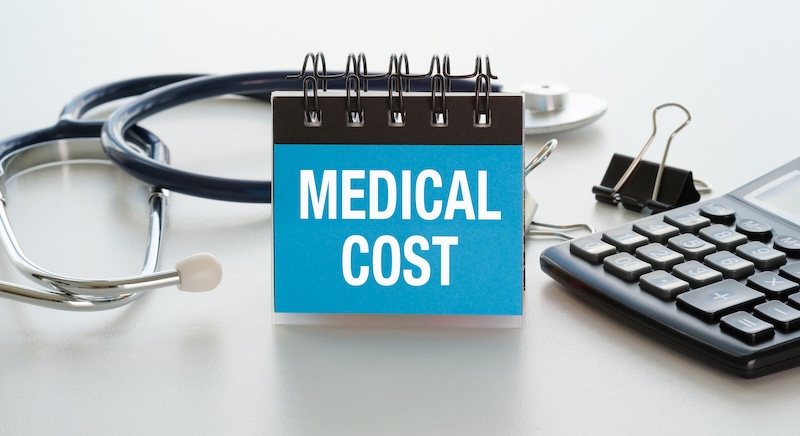 Medical cost sign sits beside stethoscope and calculator. Table surface holds office tools in clean arrangement. No people appear, no emotions shown. Ideal for budgeting, insurance