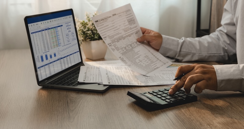 Accountant reviewing financial report and tax documents on laptop while using calculator for account analysis, real estate investment planning, expense management, and reporting workflow.